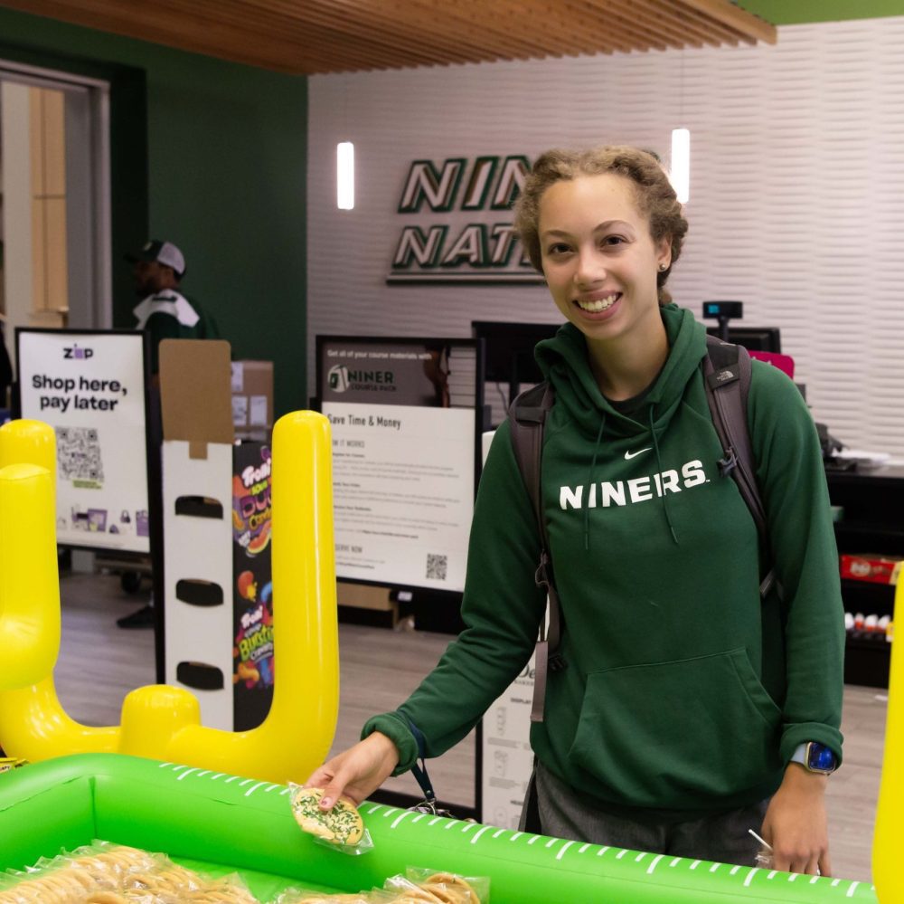 Student with green Niners sweatshirt picks up sugar cookie with green and white sprinkles from College Colors Day giveaway table in Charlotte Barnes & Noble